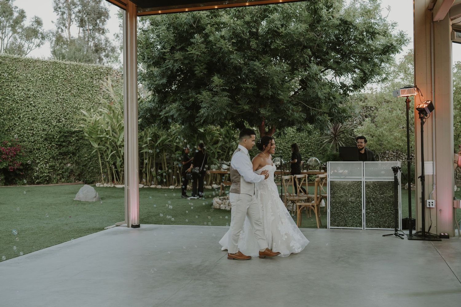 Newlyweds share their first dance on outdoor patio surrounded by lush greenery at wedding reception.