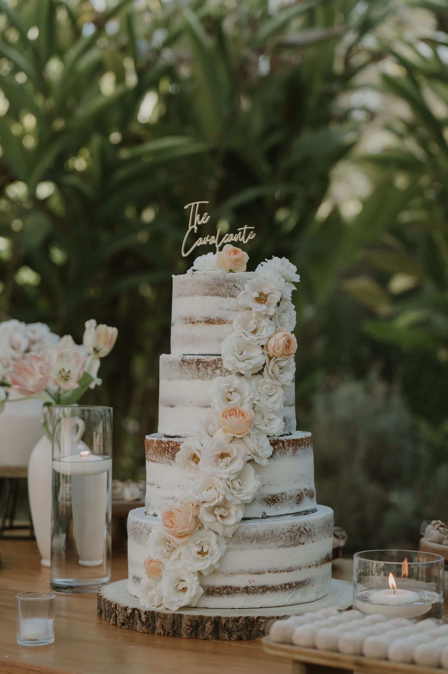 Three-tier semi-naked wedding cake with white and blush roses, gold topper, displayed on wood slice outdoors.
