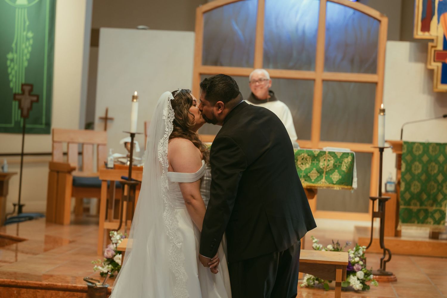 Bride and groom share first kiss at church altar during wedding ceremony with officiant.