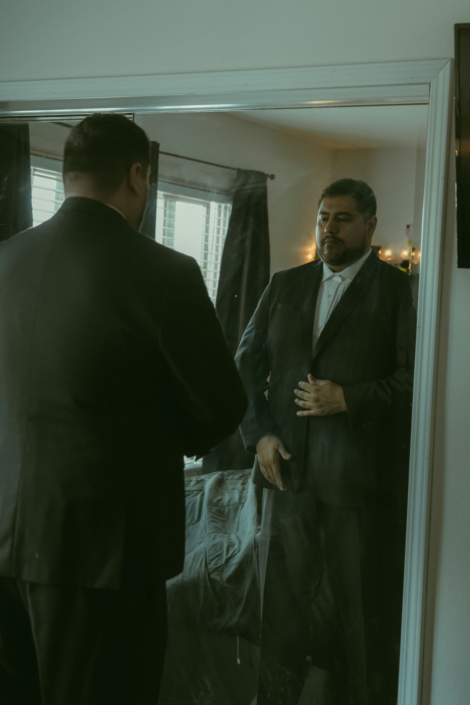 A bearded man in a dark suit adjusts his jacket while standing in front of a bedroom mirror.