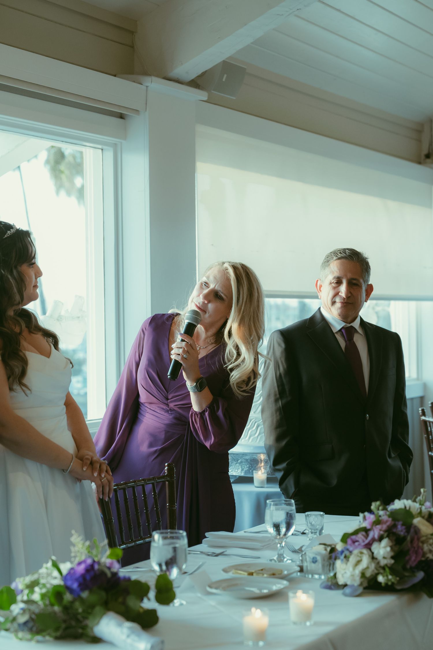 Blonde woman in purple dress gives a toast with microphone at an elegant wedding reception dinner.