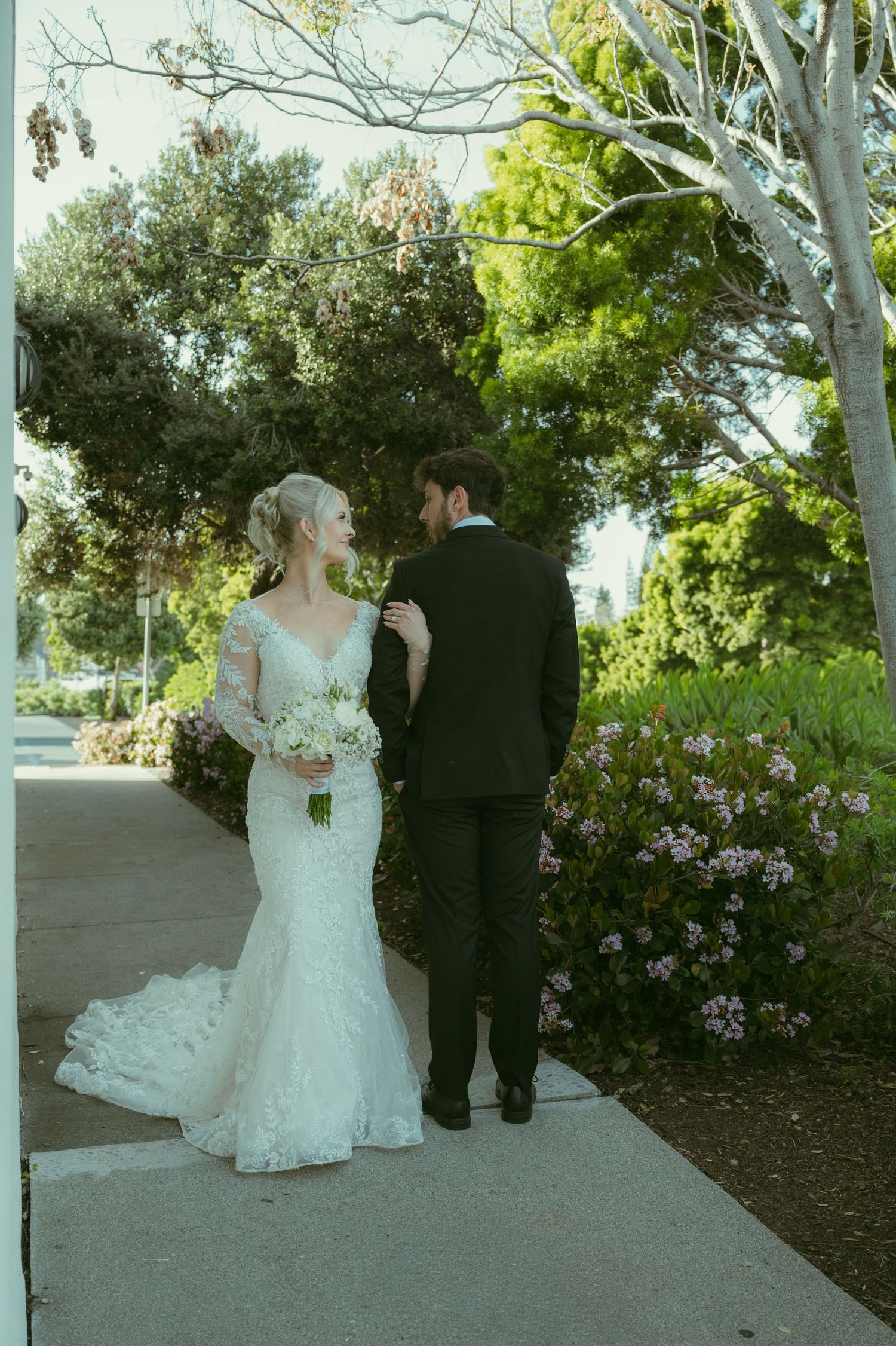 Bride in lace mermaid gown holding white bouquet walks with groom in black suit along garden path.