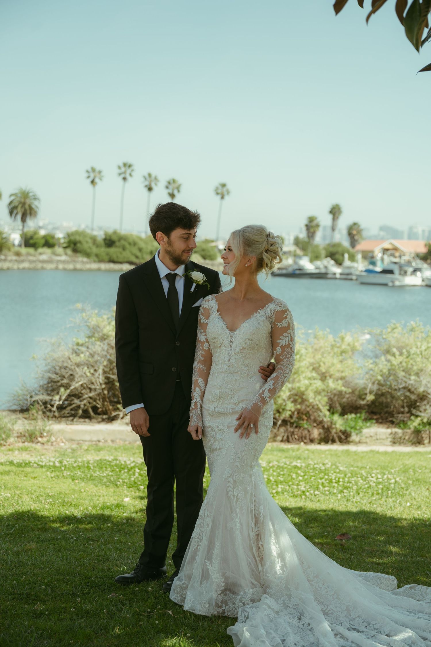 Bride in lace mermaid gown and groom in black suit share a romantic glance by a waterfront with palm trees.