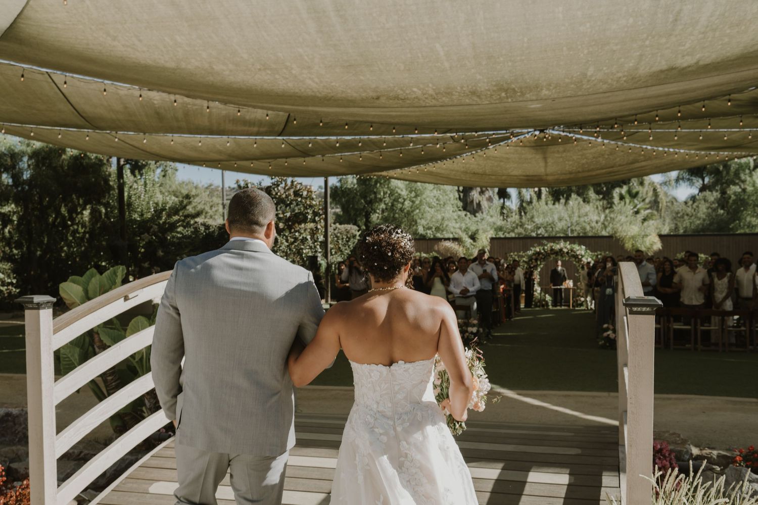 Bride and groom walking hand in hand toward outdoor wedding ceremony under elegant draped canopy.