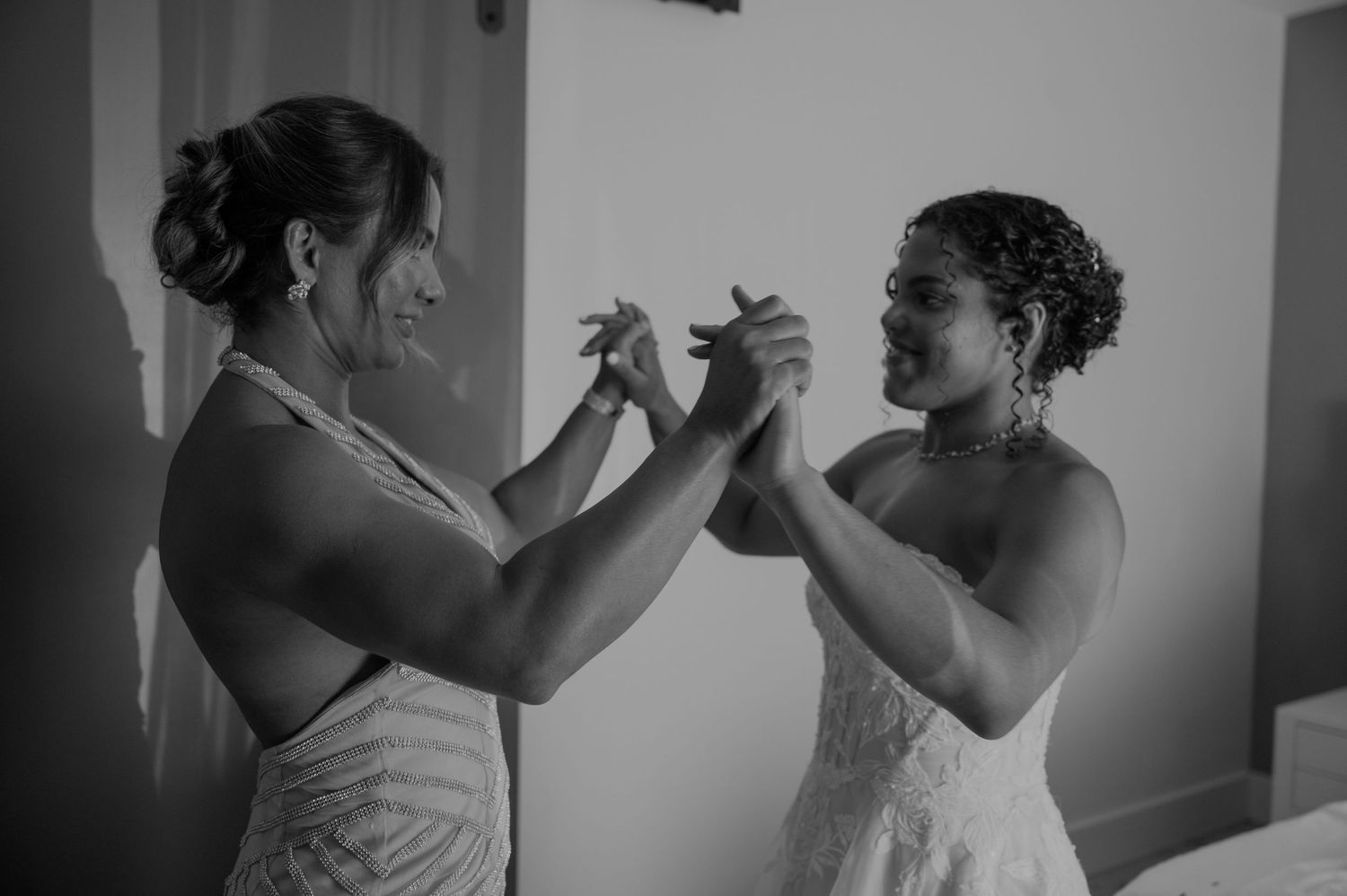 Two women in formal attire hold hands and laugh together during a wedding preparation moment.