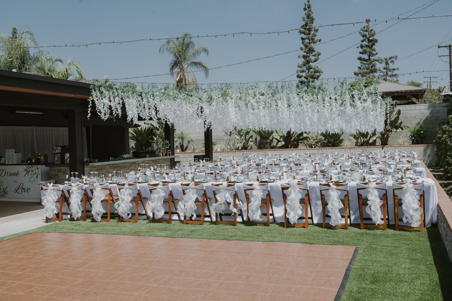 Outdoor wedding reception setup with white chair sashes, gift bags, and floral arch backdrop on artificial turf dance floor.