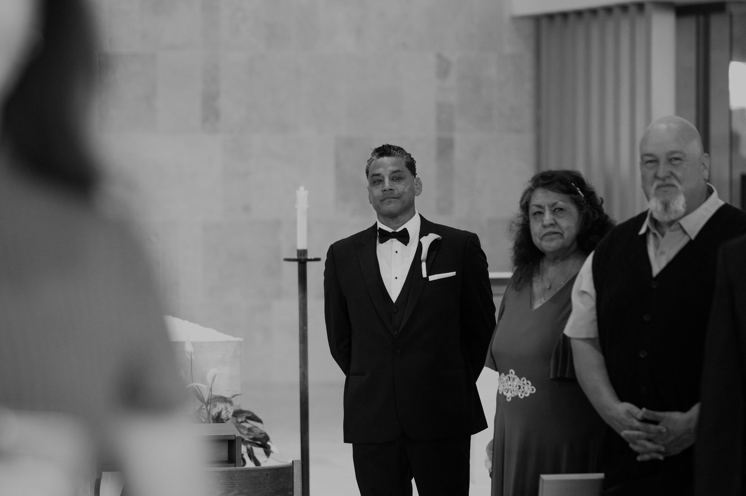 Groom in tuxedo stands at church altar with family members during wedding ceremony, black and white photo.