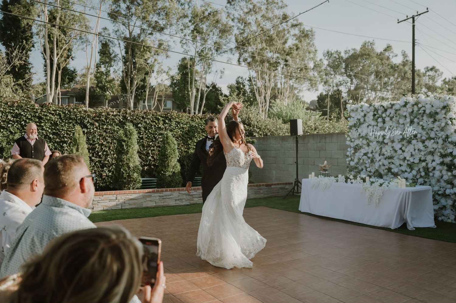 Bride and groom share first dance on outdoor dance floor surrounded by guests and floral wall.