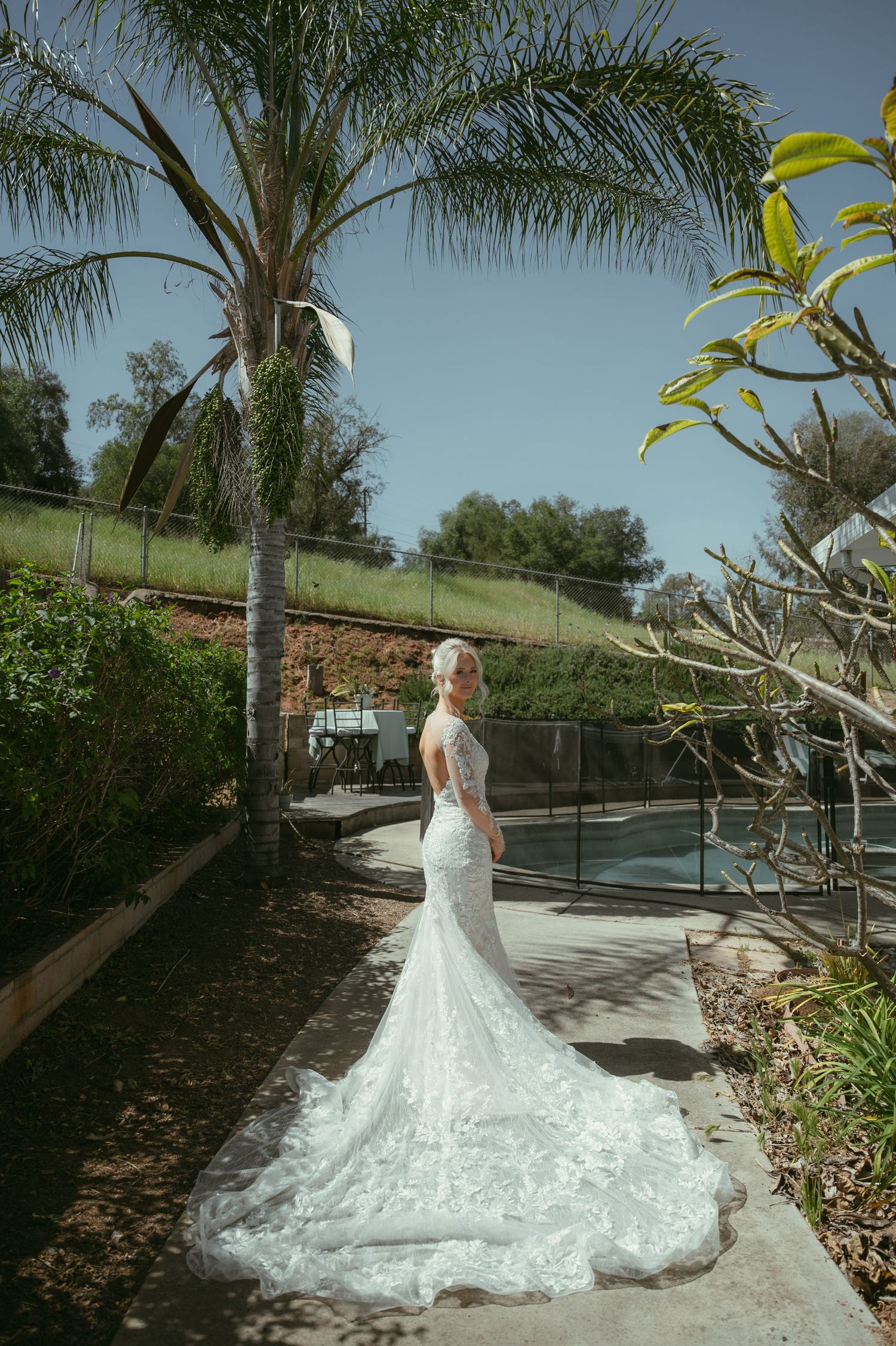 Elegant bride in a lace mermaid wedding gown standing near a palm tree by a pool.