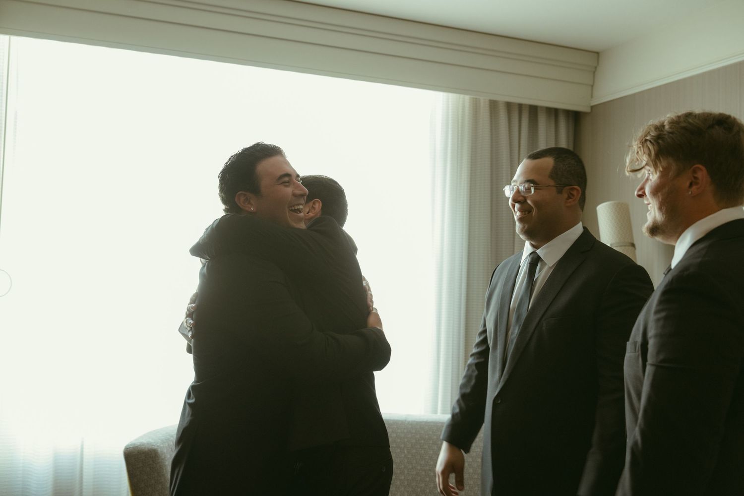 Groom sharing an emotional hug with guest while groomsmen stand nearby in hotel room.