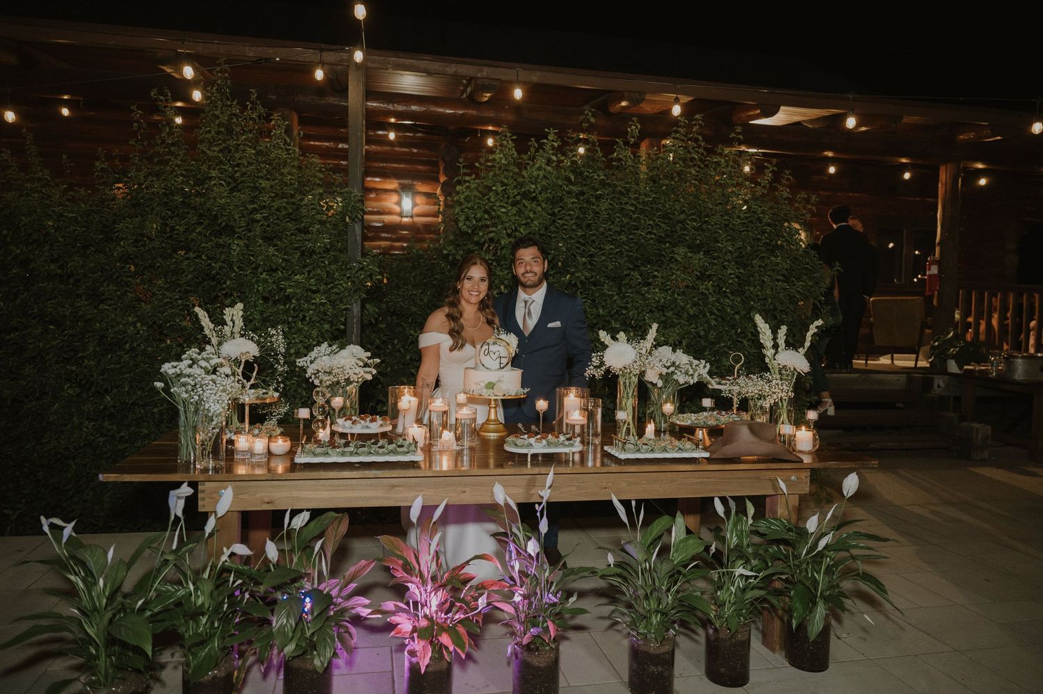 Bride and groom stand behind elegant candlelit reception table with floral arrangements and potted plants at outdoor wedding.