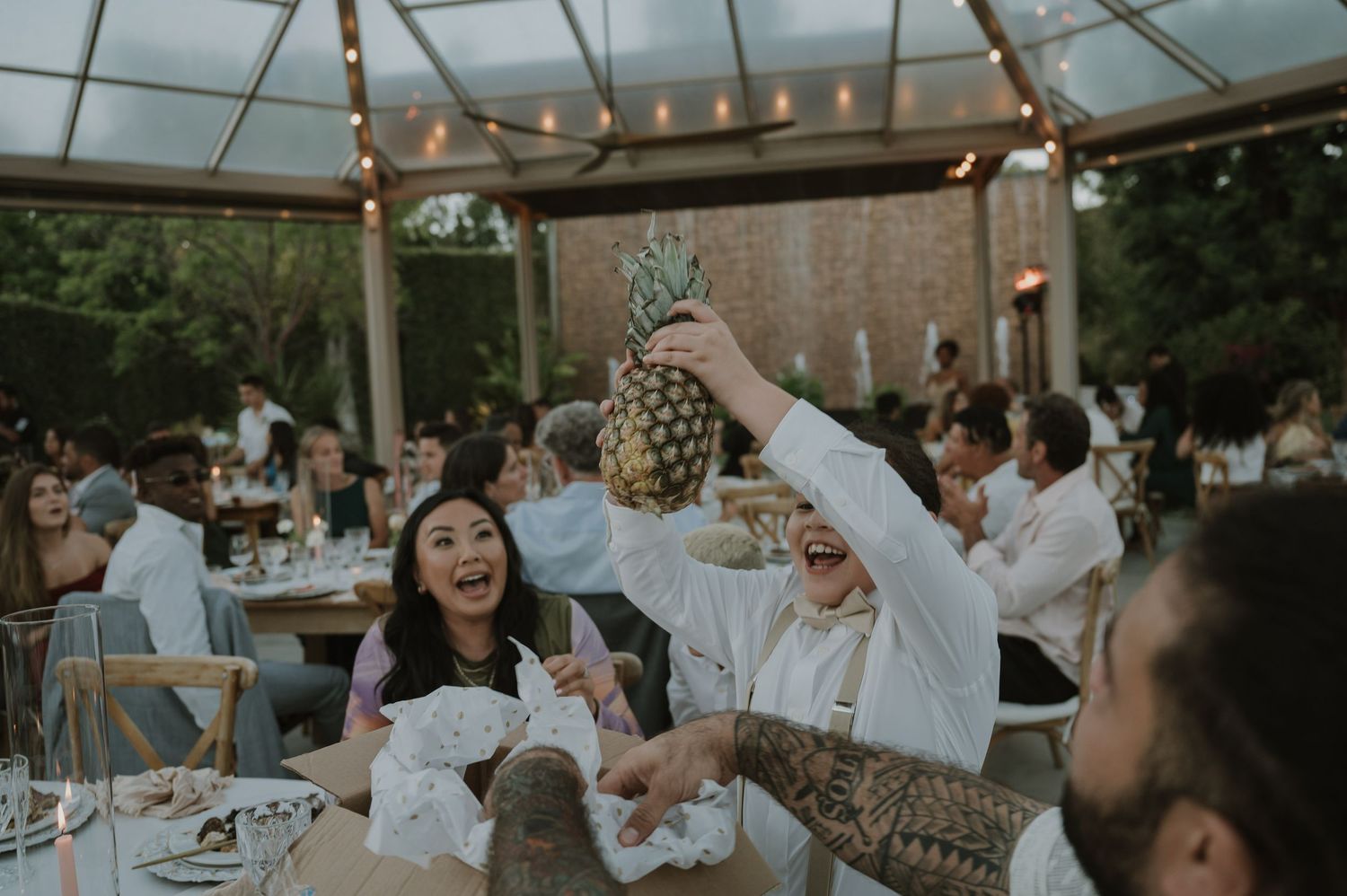 Wedding guest triumphantly holds up a pineapple at elegant outdoor reception dinner under string lights.