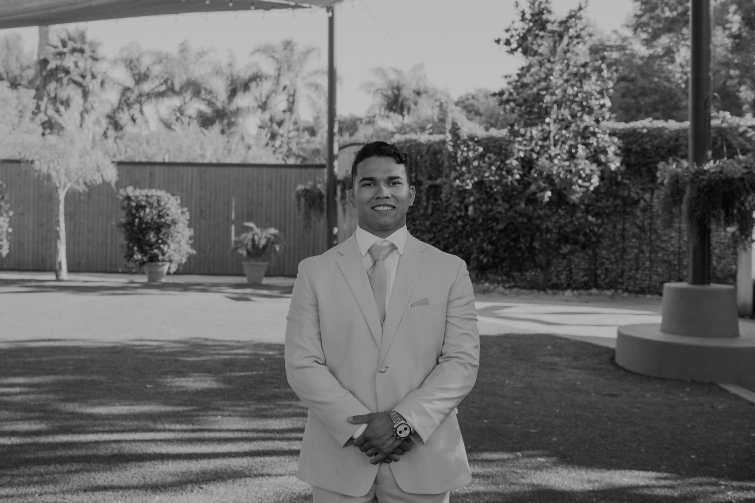 Man in light suit smiling outdoors at a wedding venue surrounded by palm trees and greenery.
