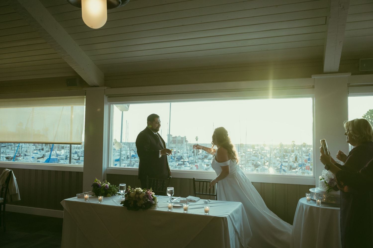 Bride and groom share a moment at sweetheart table with stunning sunset harbor view through venue windows.