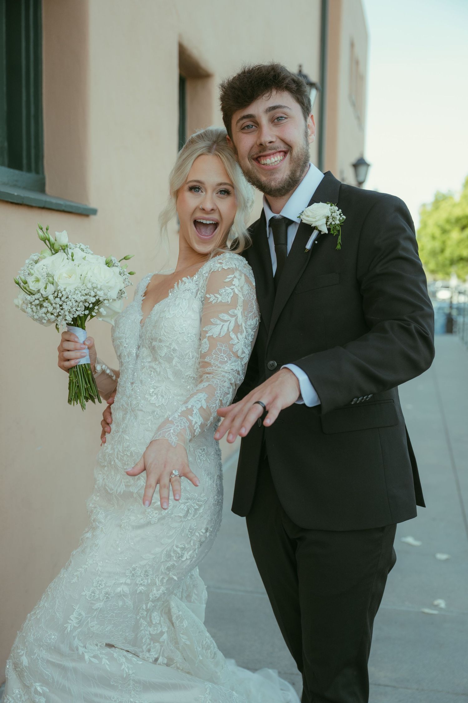 Joyful newlyweds pose outdoors, bride in lace gown holding white bouquet, groom in black suit, both showing wedding rings.