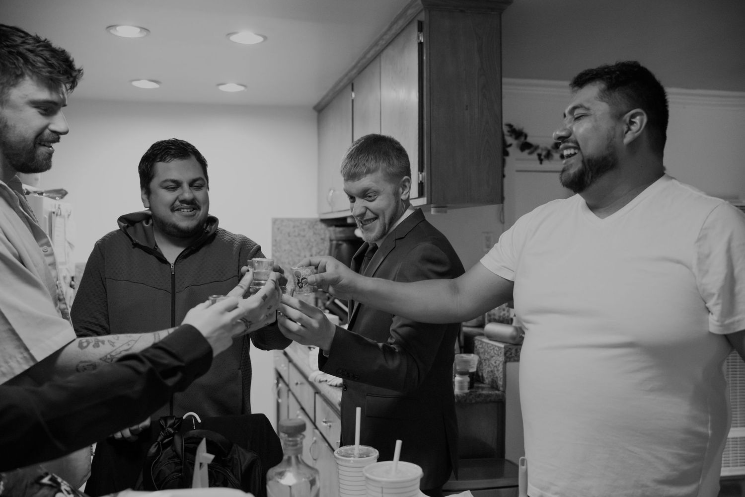 Four men laughing and toasting drinks together in a kitchen in a black and white photo.