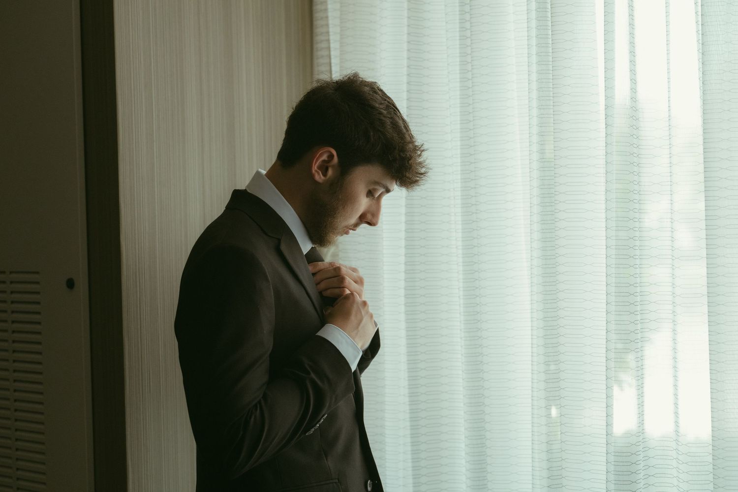 Dapper groom in a dark suit adjusting his tie near sheer curtains on his wedding day.