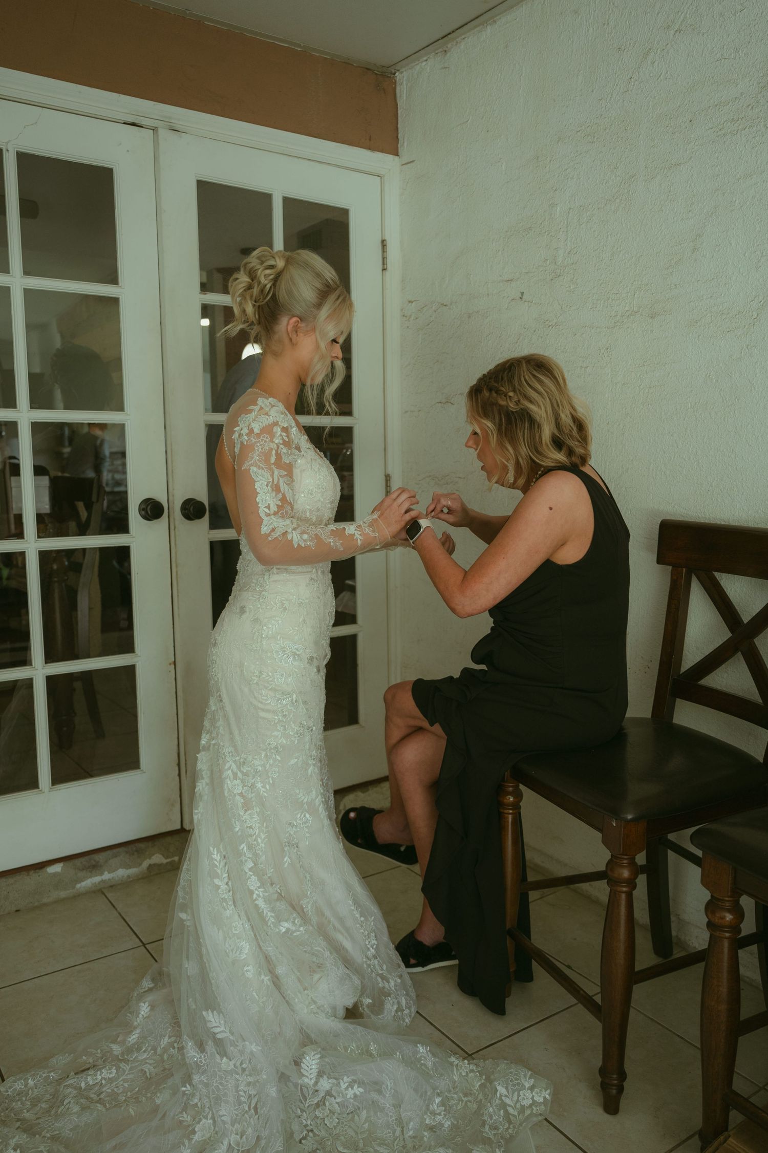 A bride in a lace wedding gown stands as a woman in black adjusts her dress near French doors.