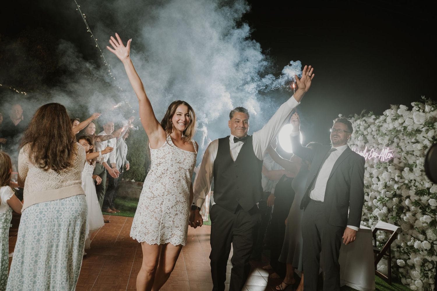 Joyful bride and groom celebrate wedding exit with raised hands, sparklers, and smoke effects at night.