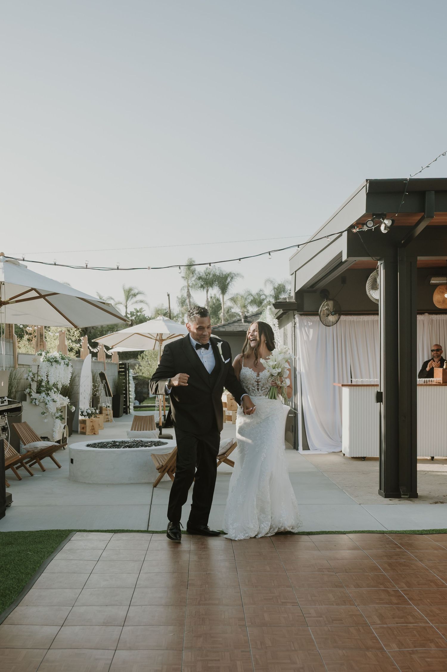 Bride and groom make grand entrance at outdoor wedding reception with string lights and palm trees.