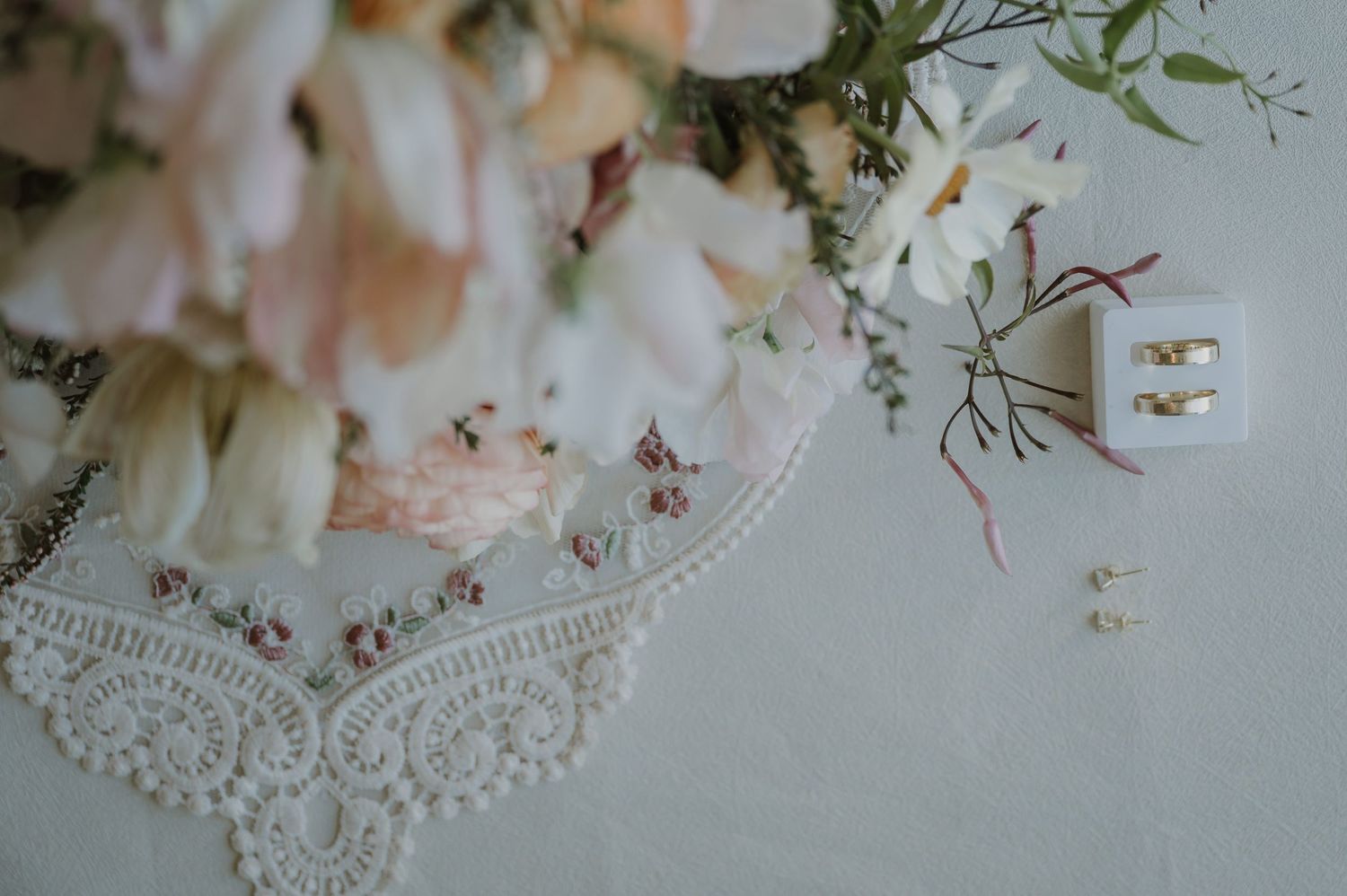Elegant wedding flat lay with blush floral bouquet, lace handkerchief, and gold earrings on white background.