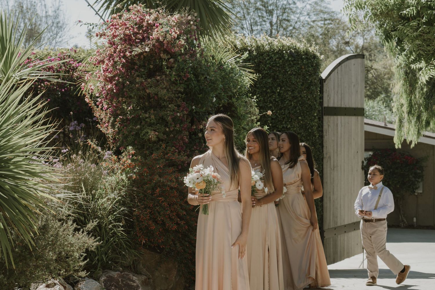 Bridesmaids in blush gowns walk with bouquets through a lush garden setting on a sunny wedding day.