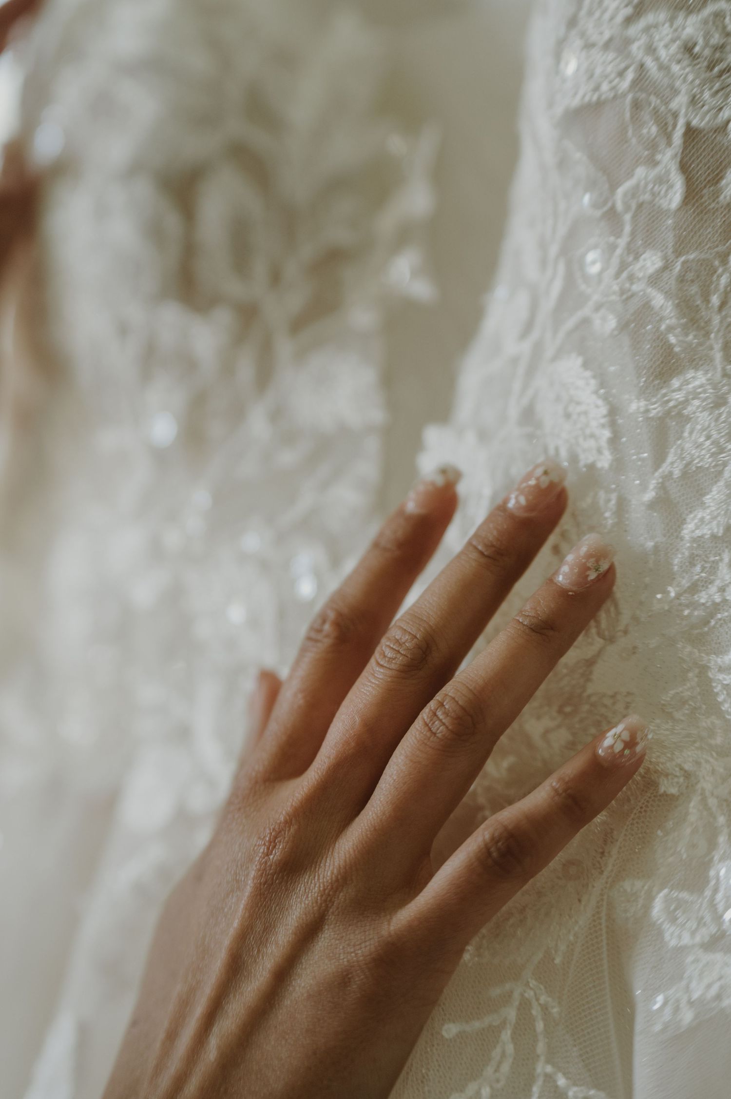 A bride's hand with delicate nail art gently touches intricate white lace fabric of a wedding dress.