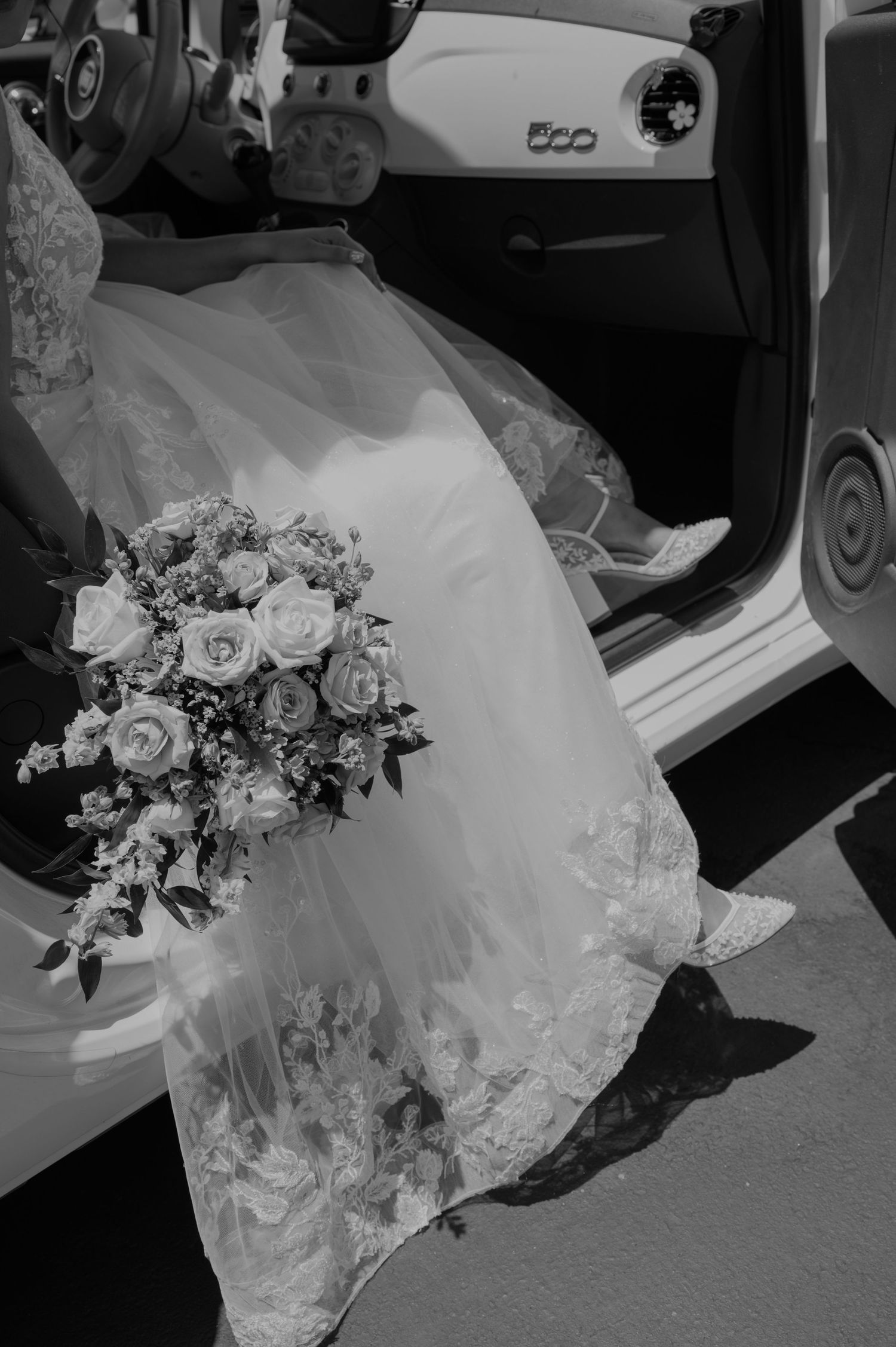 Black and white photo of a bride in a lace gown holding a rose bouquet while stepping out of a white car.