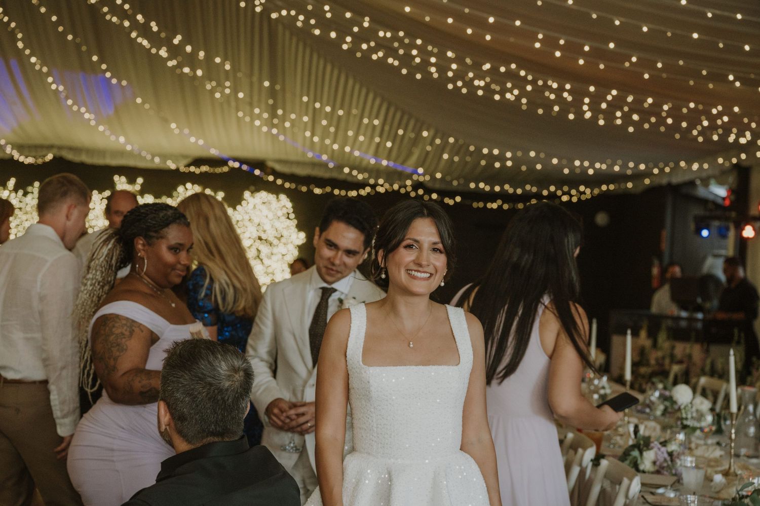 Smiling bride in white dress mingles with guests under romantic fairy light canopy at wedding reception.