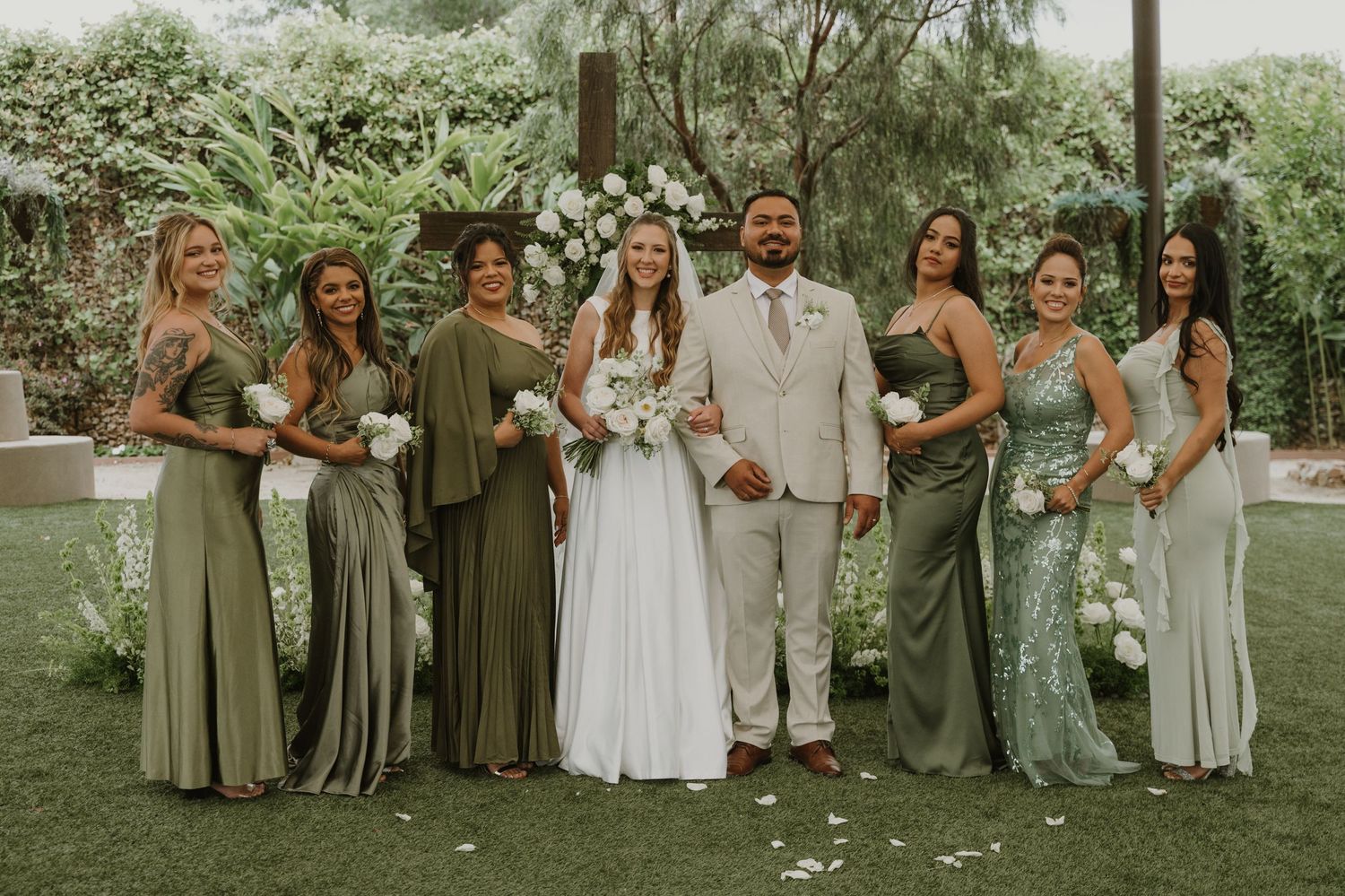 Wedding party poses outdoors with bride and groom surrounded by bridesmaids in sage green dresses holding white bouquets.