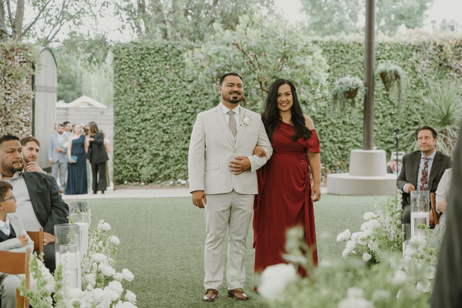 Groom in light suit walks down outdoor wedding aisle with woman in red dress, surrounded by white floral arrangements.