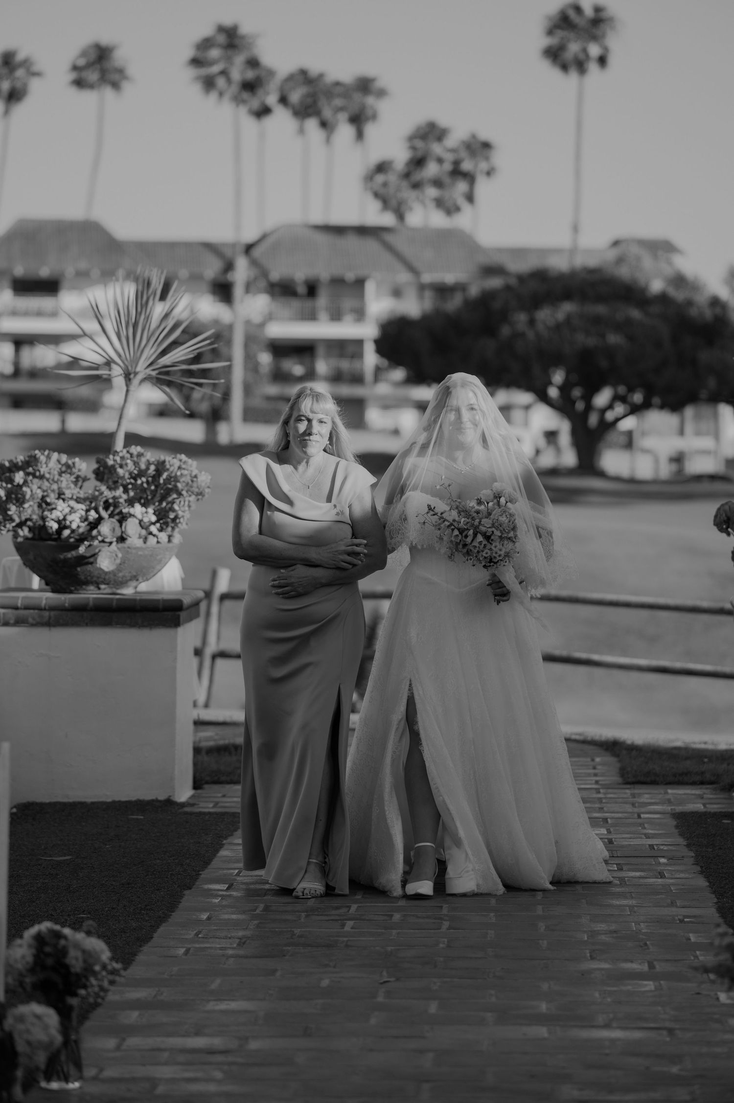 Bride in white gown and veil walks down aisle with bridesmaid in elegant gown at outdoor wedding ceremony.