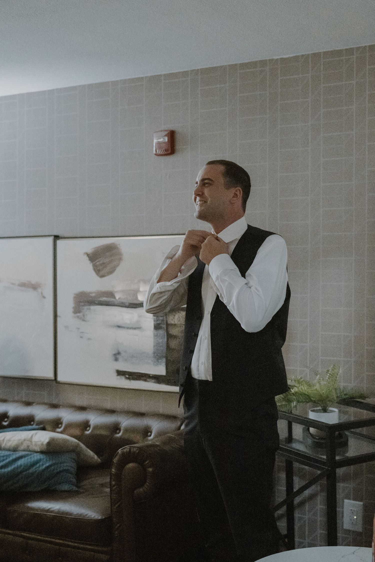 A smiling groom in a black vest and white shirt adjusts his tie while getting ready in an elegant hotel lobby.