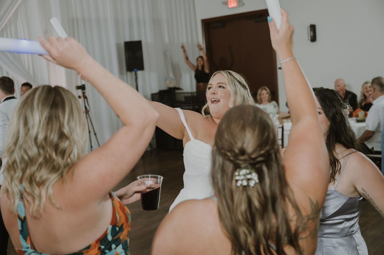 Bride in white bodysuit laughs and dances with guests at a lively wedding reception celebration.