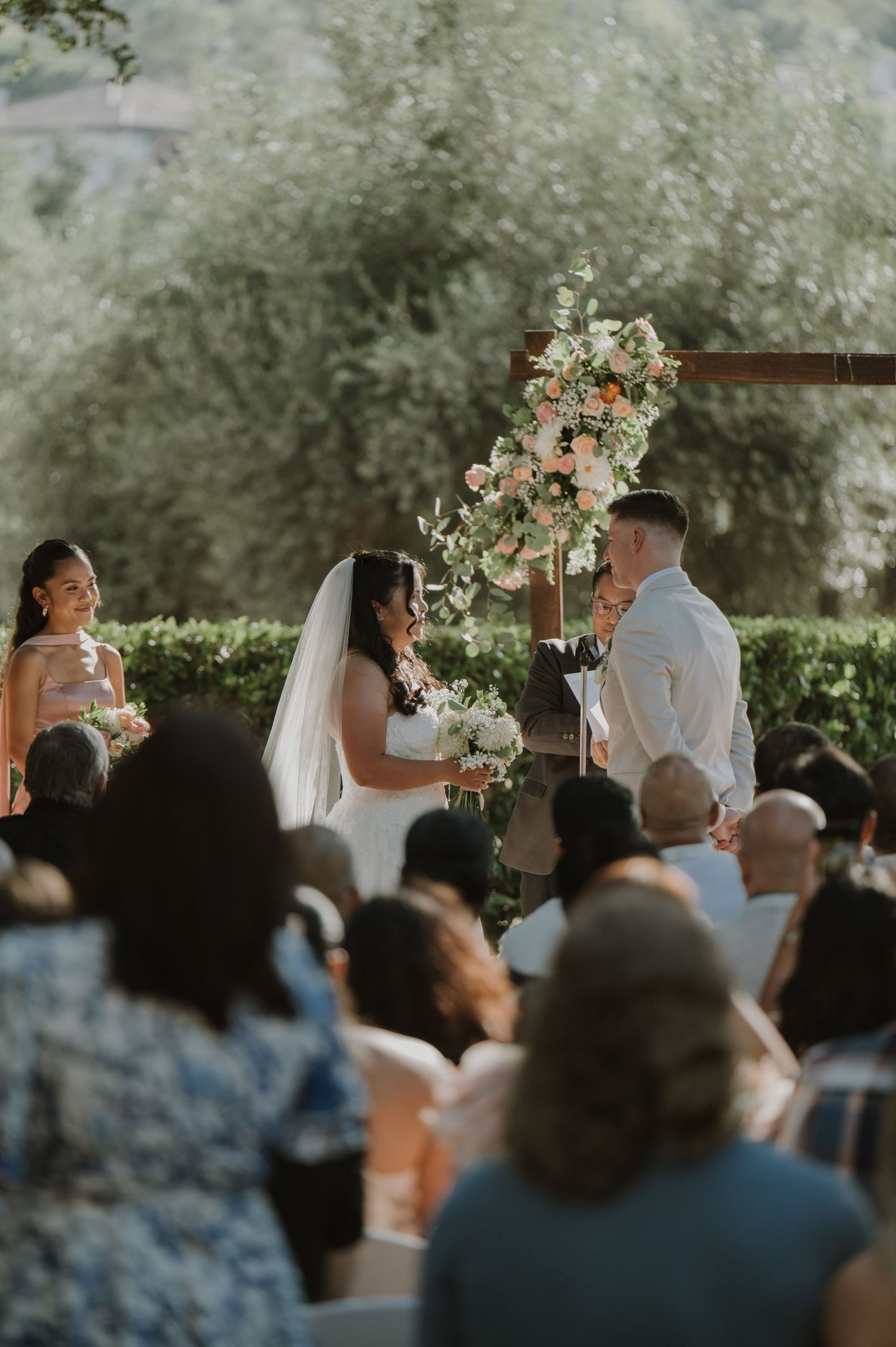 Outdoor wedding ceremony with bride and groom at floral arch, guests seated, surrounded by lush greenery.