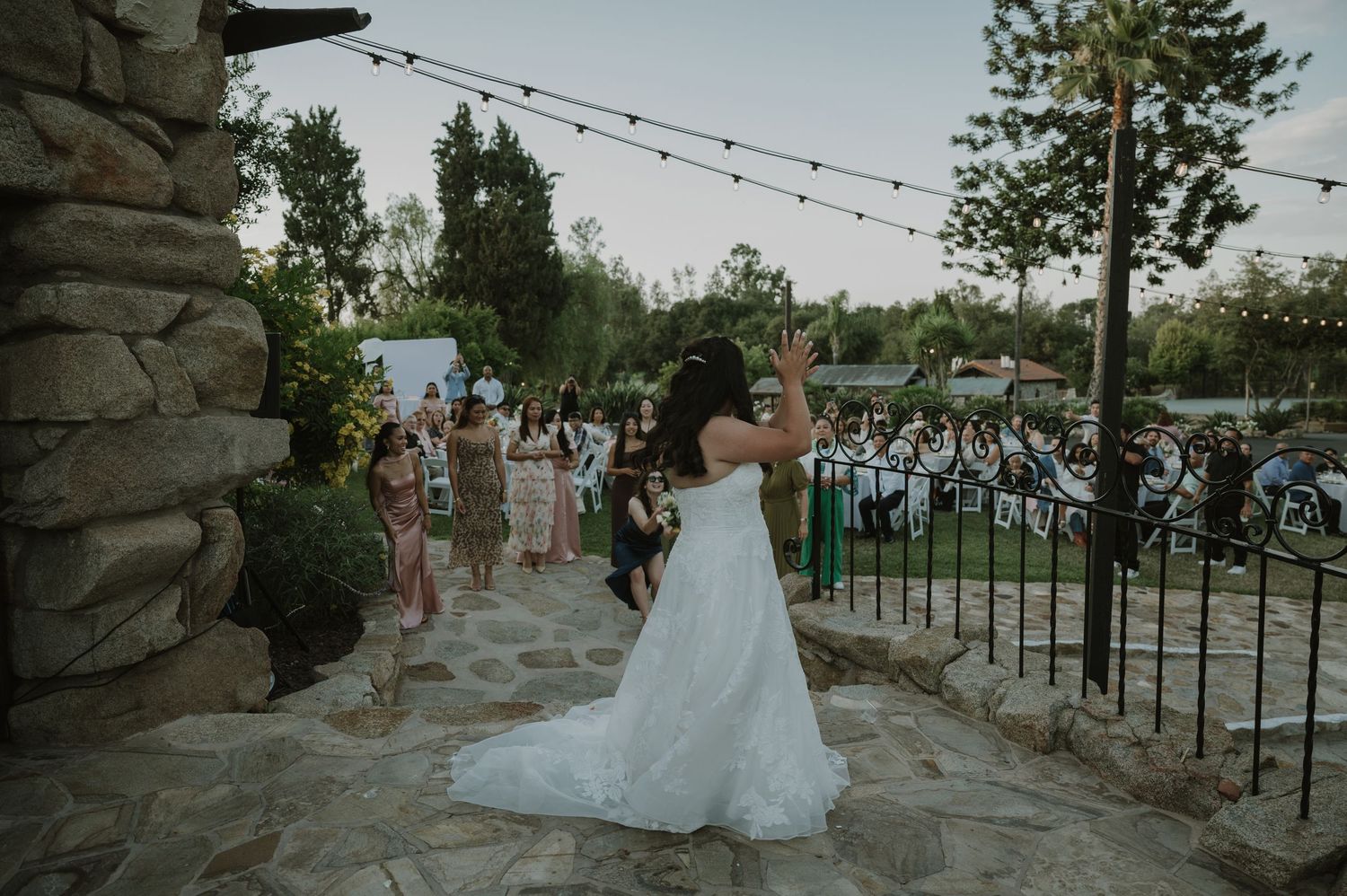 Bride in elegant white ball gown throwing bouquet to crowd at outdoor evening wedding celebration.