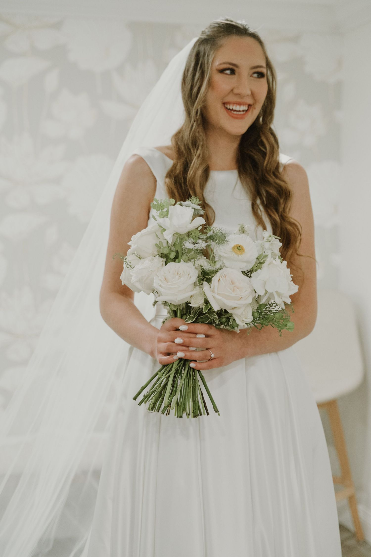 Smiling bride in white gown and veil holding elegant white rose bouquet on her wedding day.