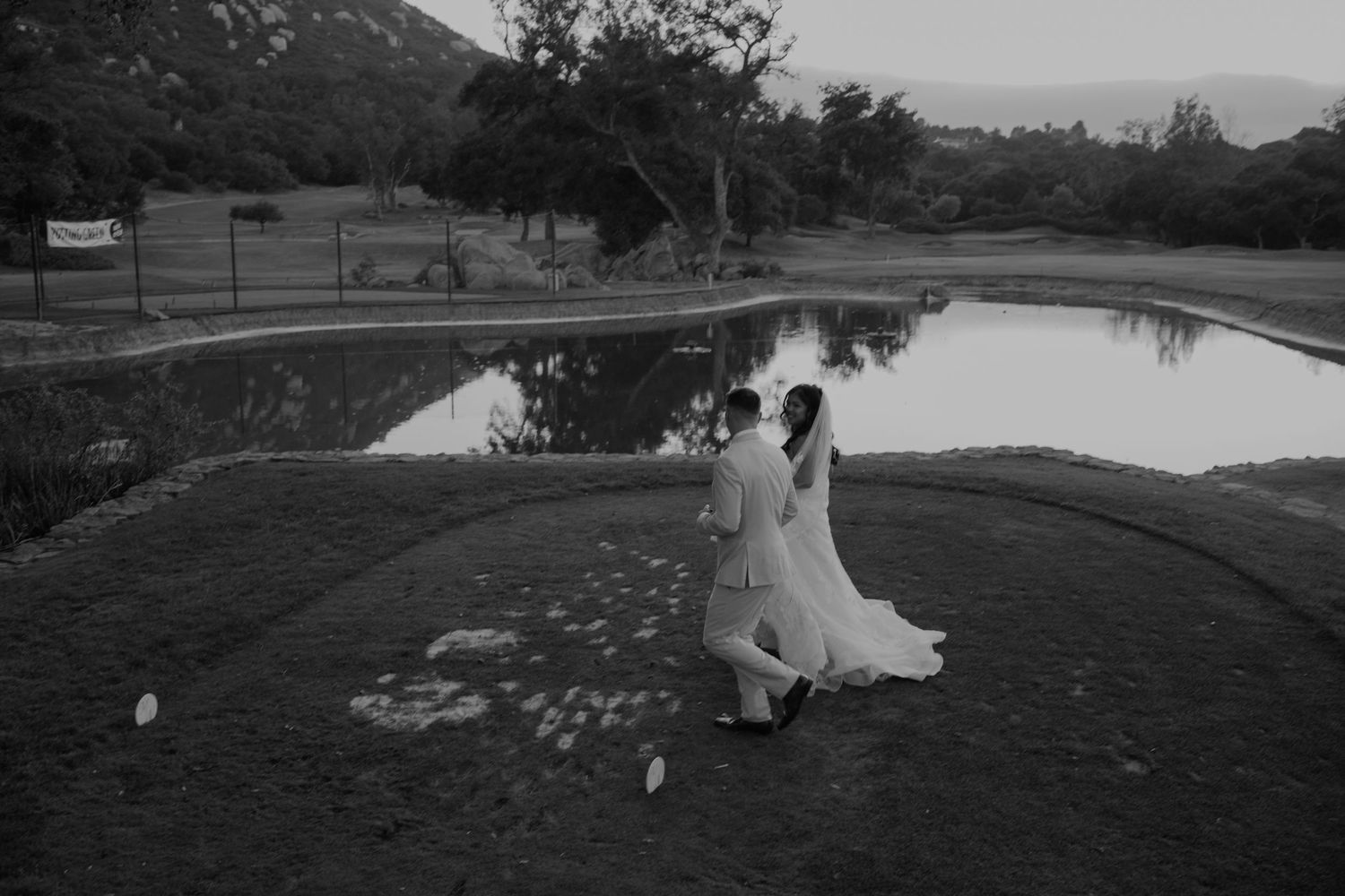 Black and white photo of couple in wedding attire by reflective pond at sunset.