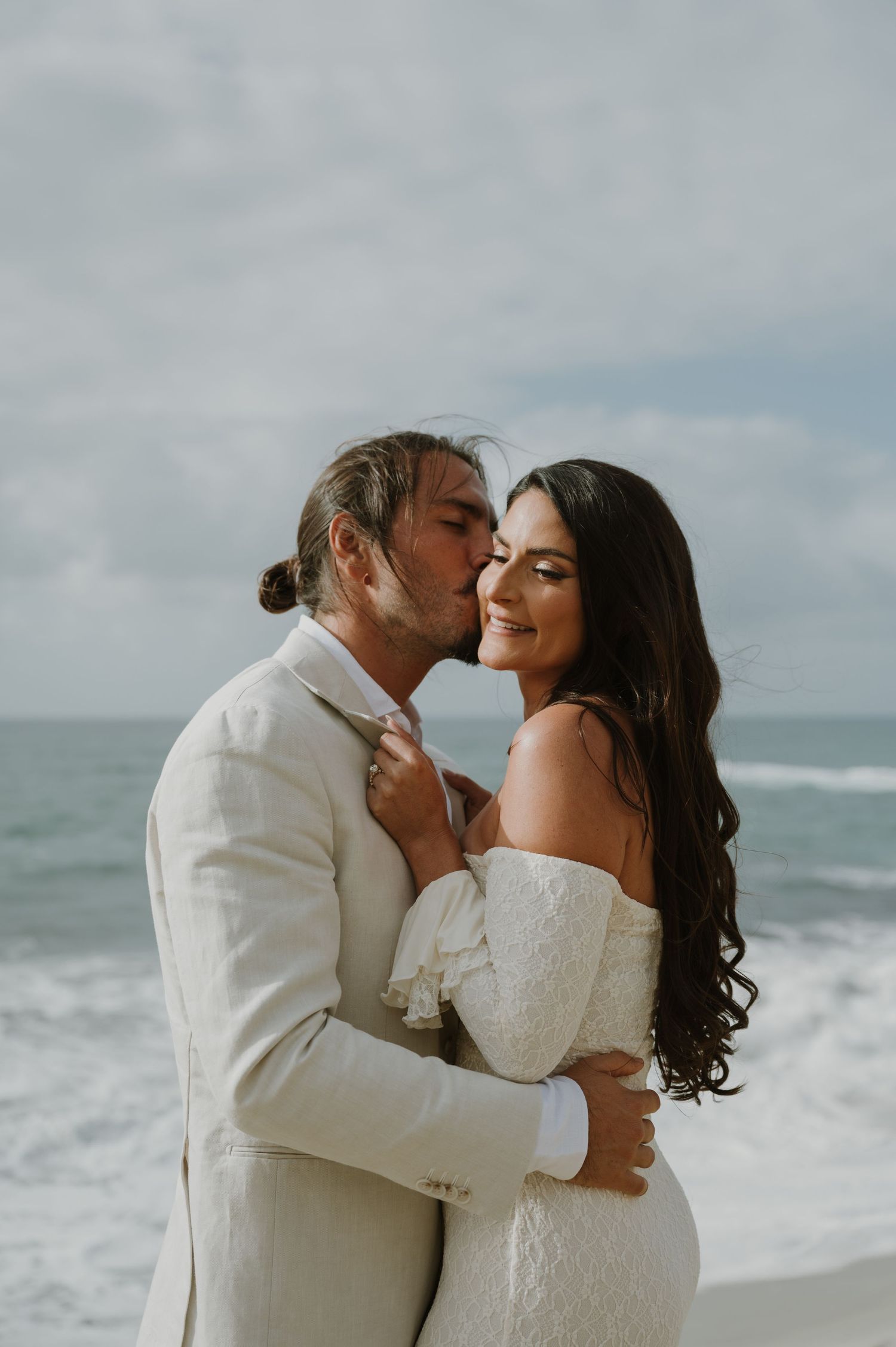 A couple in white wedding attire shares tender moments on a beach with crashing waves.