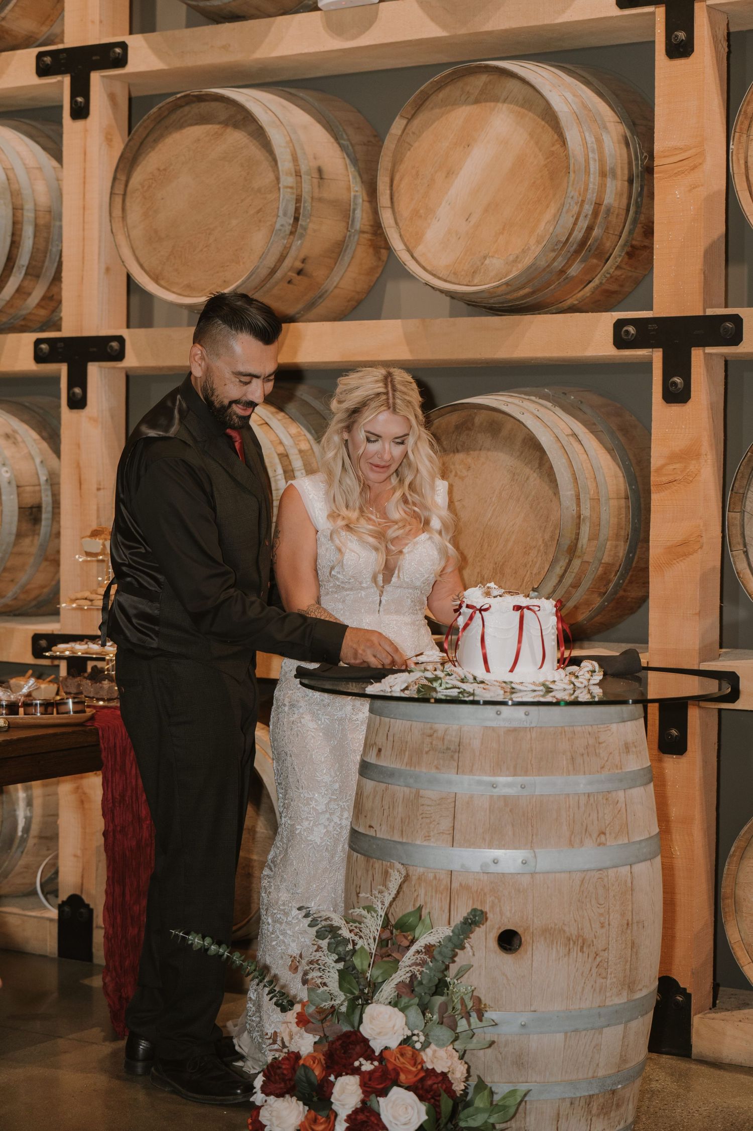 Wedding cake cutting ceremony in front of wooden wine barrels in a rustic winery setting.