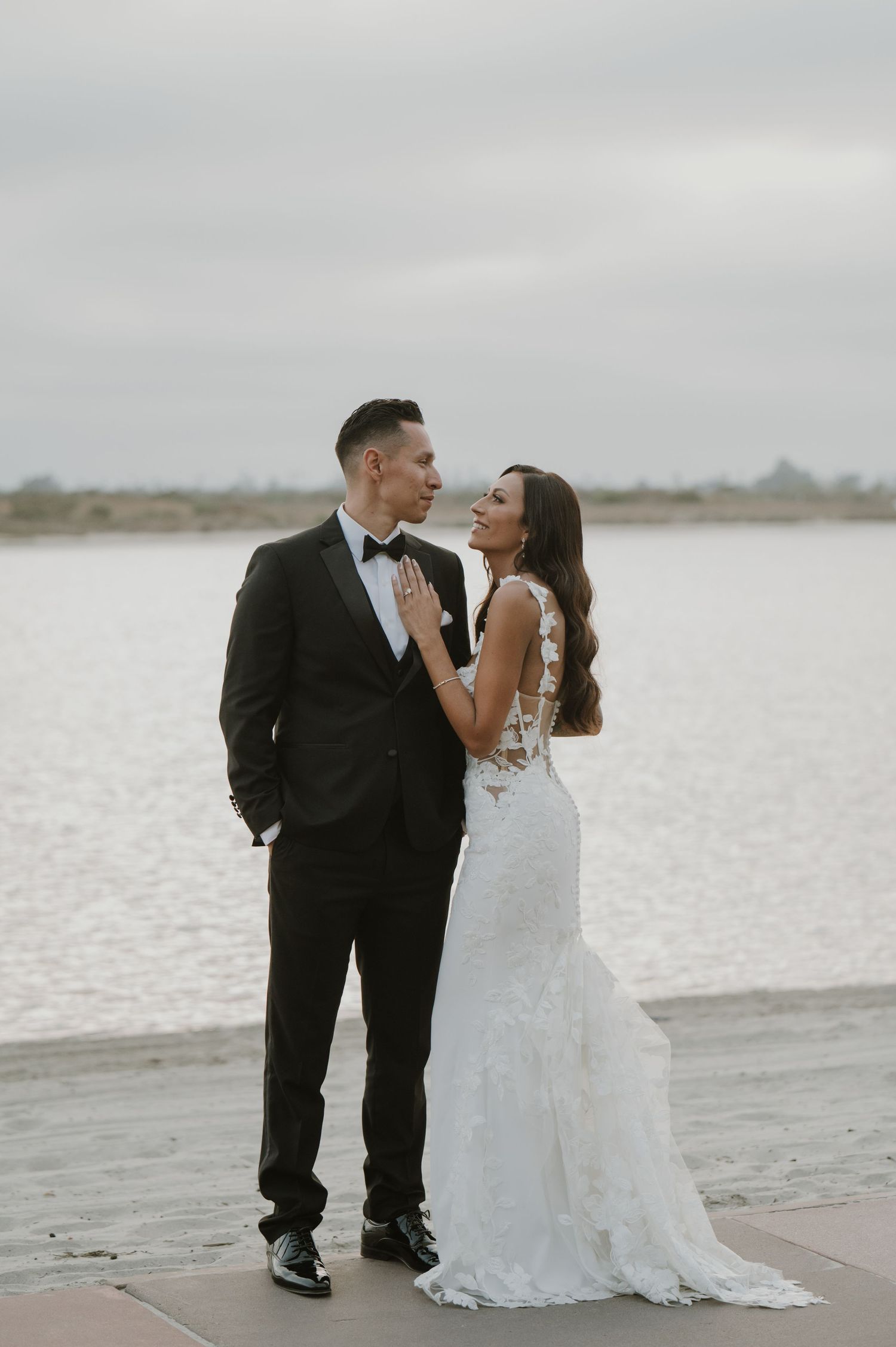A romantic wedding couple shares a tender moment on a sandy beach with a cloudy sky backdrop.