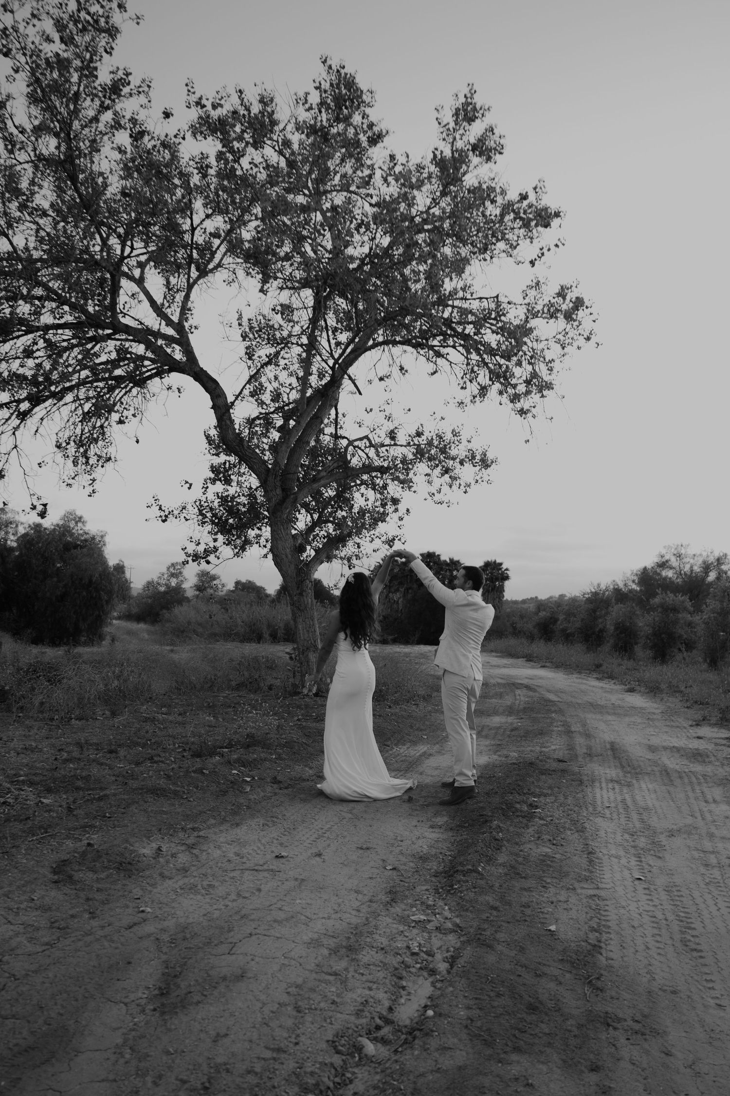 Black and white photo of a couple dancing on a rural dirt road beside a tree at dusk.