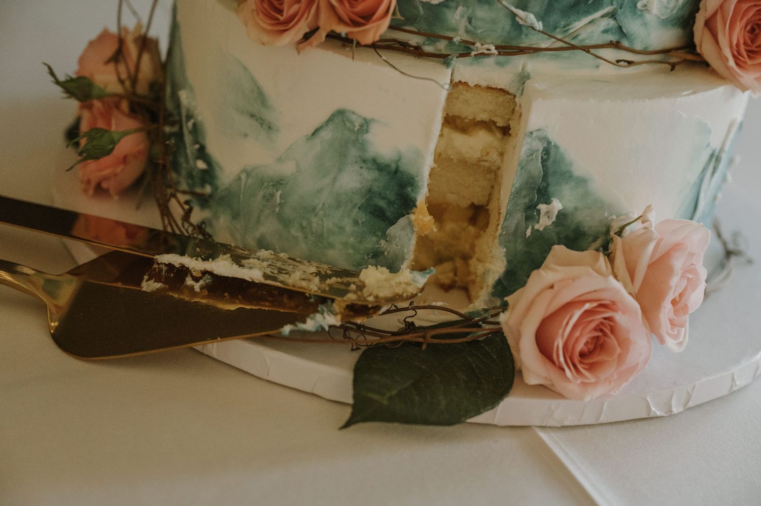 Elegant two-tier wedding cake with watercolor mountain design and pink roses being cut with a gold server.