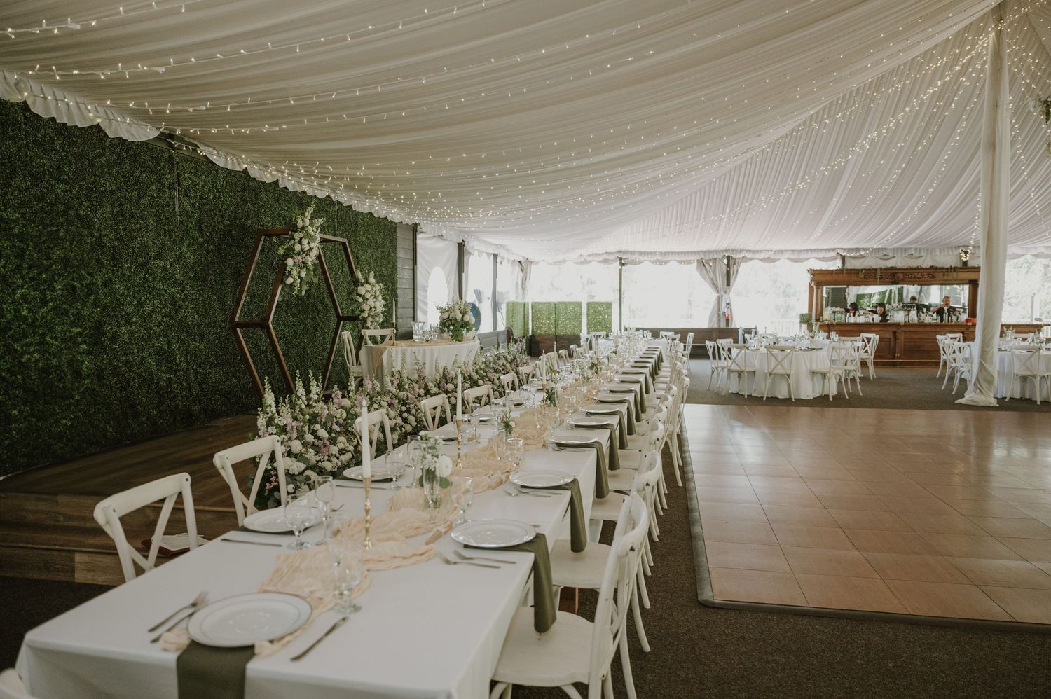Elegant wedding reception tent with long white tables, cross-back chairs, greenery wall backdrop, and draped ceiling lights.