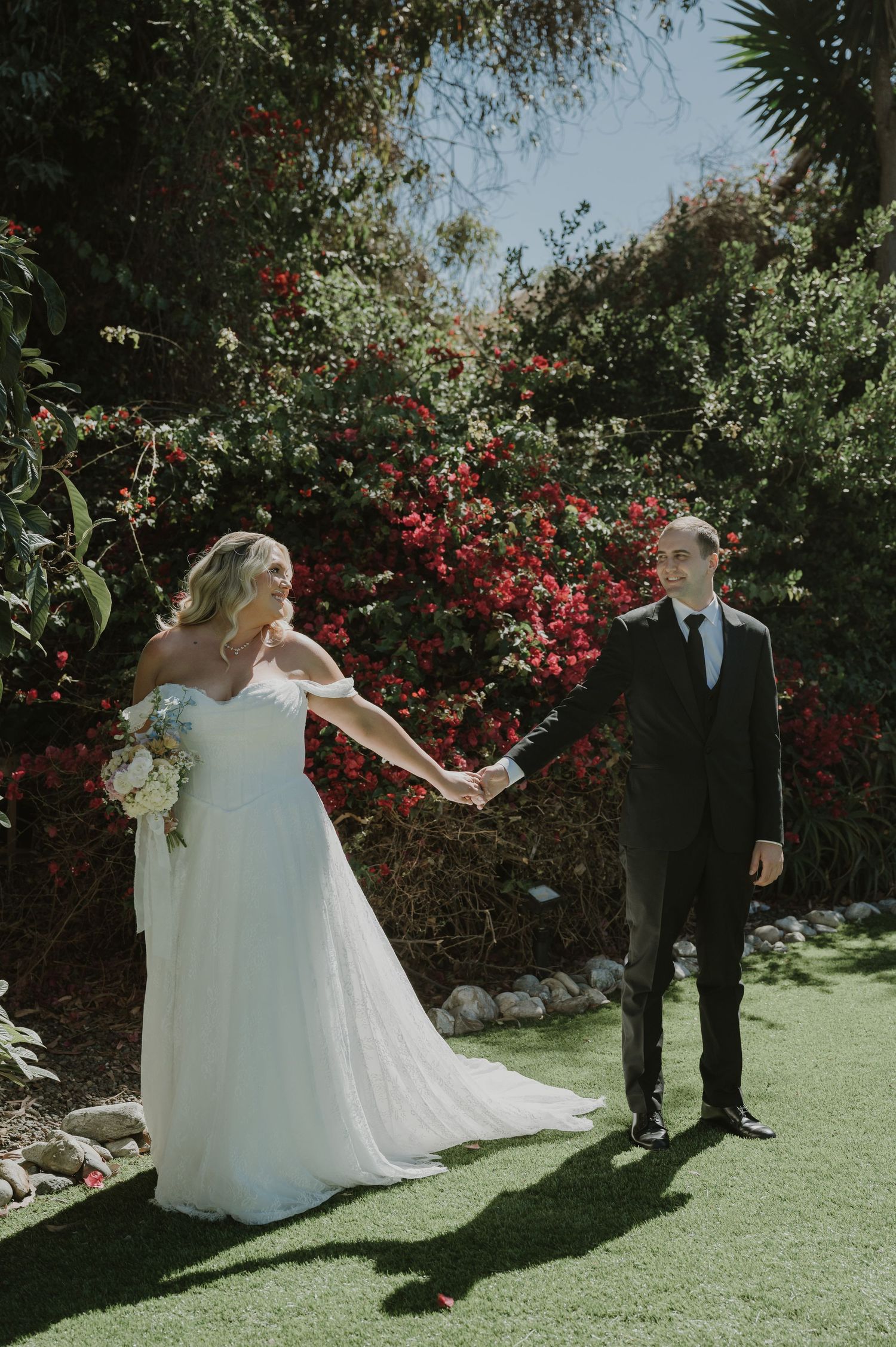 Bride and groom hold hands in a lush garden with red bougainvillea, bride wearing a white off-shoulder gown holding a bouquet.