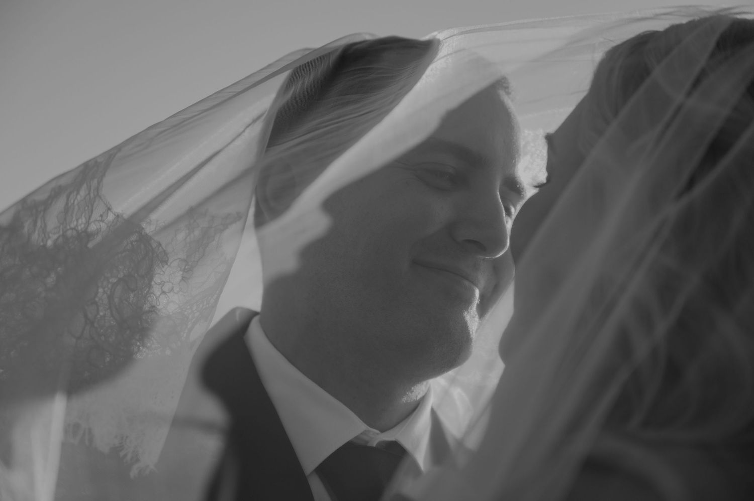 Black and white wedding photo of bride and groom sharing intimate moment under flowing veil.