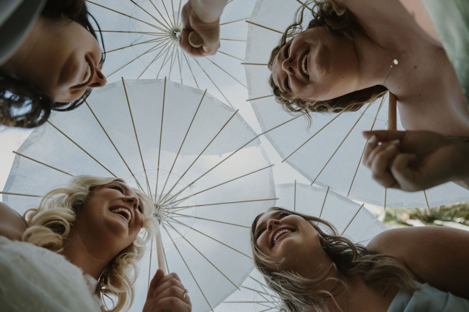 Four laughing women holding white parasols together viewed from below against a bright sky.