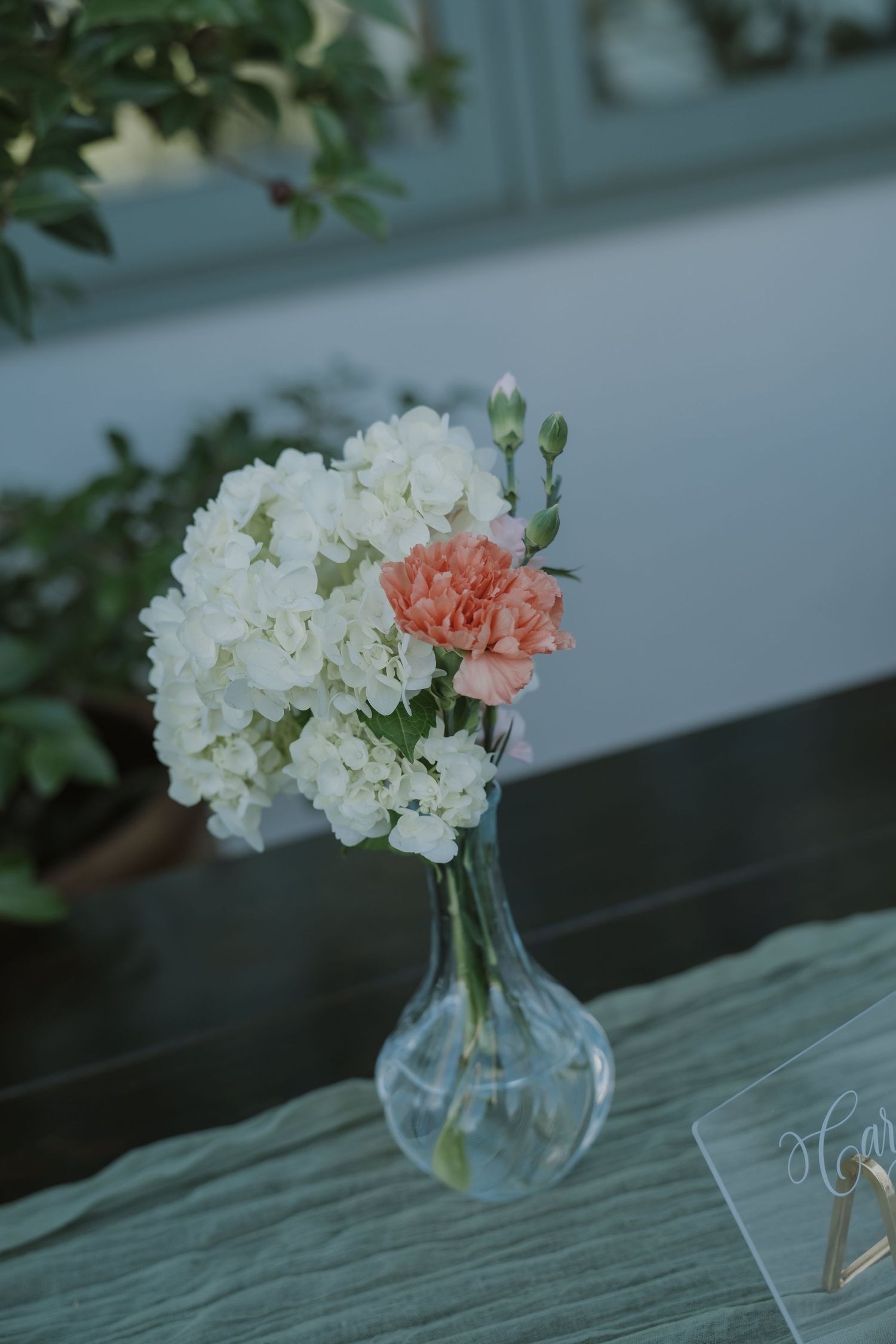 A delicate wedding centerpiece featuring white hydrangeas and a pink carnation in a clear glass bud vase on a sage green tablecloth.