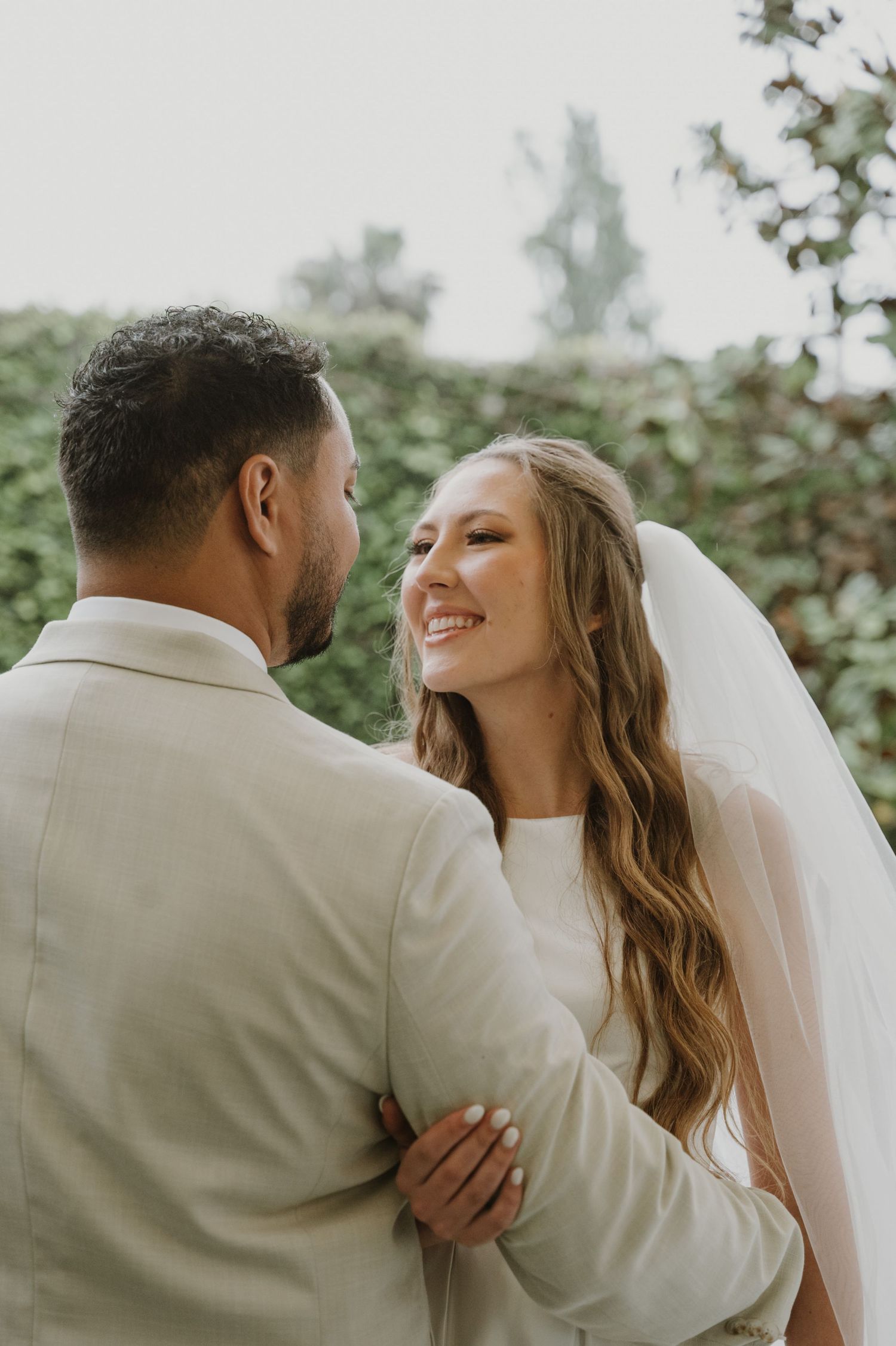 Bride in white wedding veil smiles joyfully at groom in beige suit while embracing outdoors.