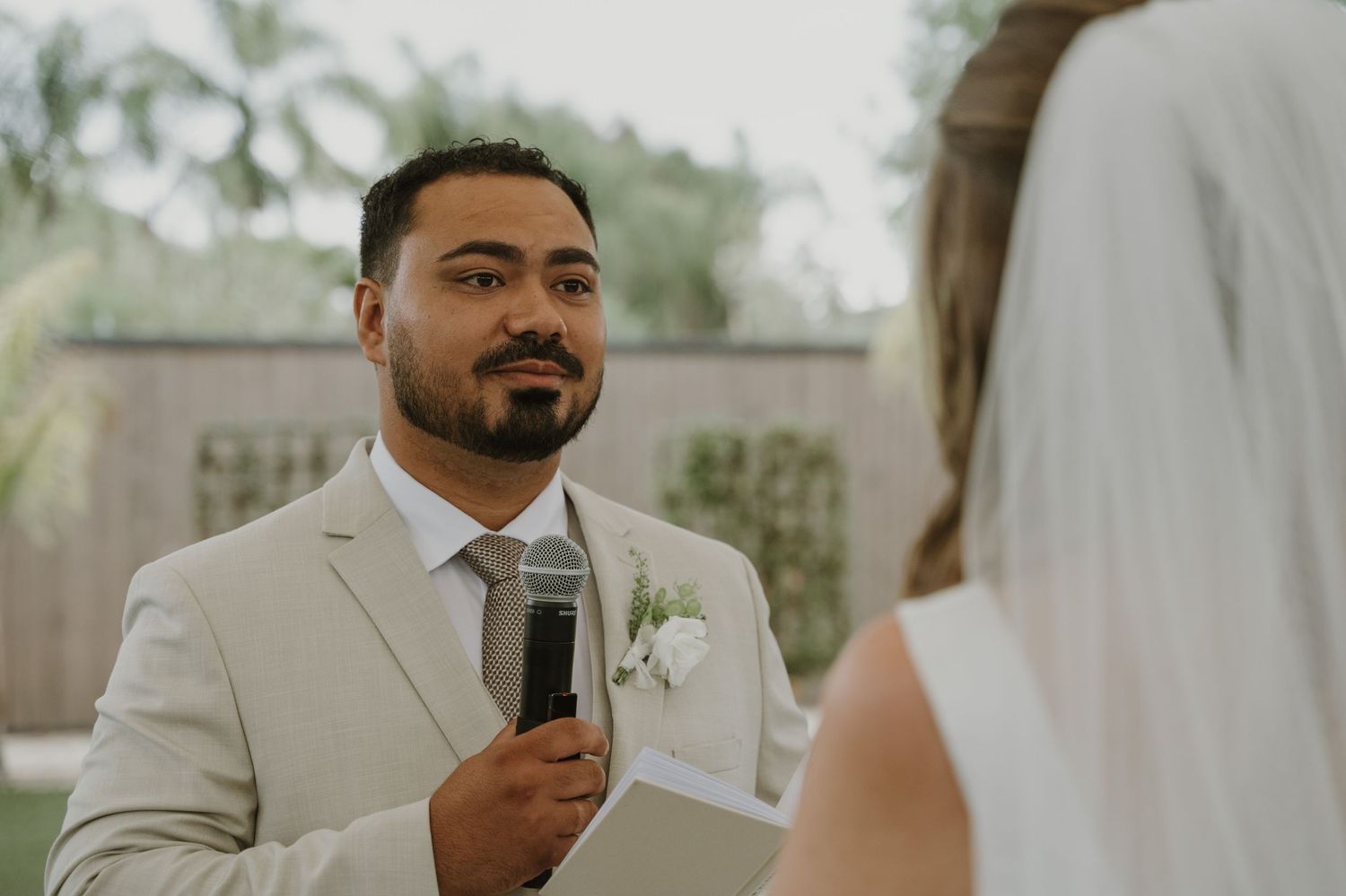 Groom in beige suit reads vows holding microphone at outdoor wedding ceremony.